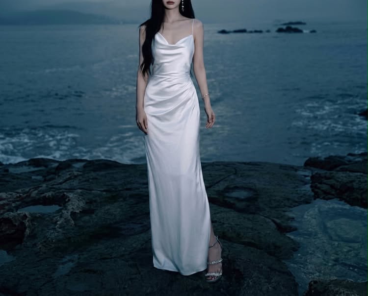 Elegant white satin cowl-neck maxi dress, styled for a formal evening event or bridal wear against a moody, scenic seaside backdrop.