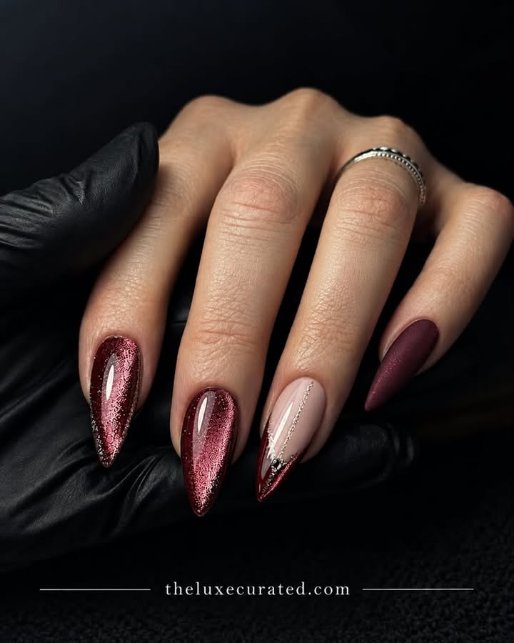 Close-up of almond-shaped burgundy cat eye nails featuring intricate foil patterns, a nude accent nail with silver glitter, and a matte dark red nail design